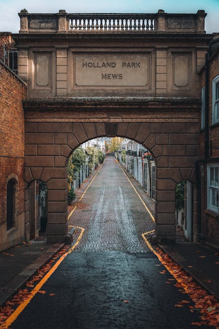A view through an arched stone gateway leading into Holland Park Mews, with a cobbled street extending beyond. The arch is constructed from reddish-brown stone blocks, featuring a rectangular inscribe