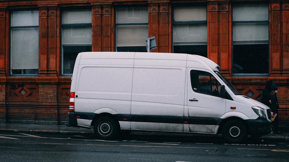 A man with a serious expression sitting inside the open back of a moving van, which is parked outside a residential property during daylight. The van's interior contains several cardboard boxes of var